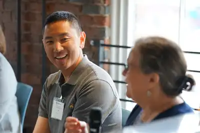 A young woman holding a drink gestures with her hand while talking to a man and a woman in a room.