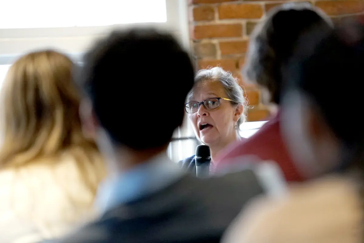 A woman in glasses holds a microphone and speaks to a crowd.