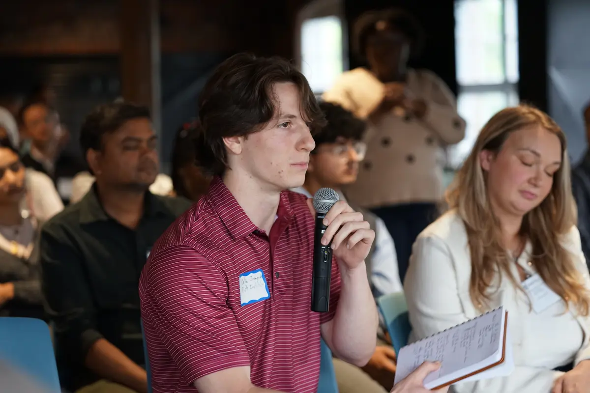 A young man in a maroon shirt holds a microphone and a notepad while speaking in a room of seated people.