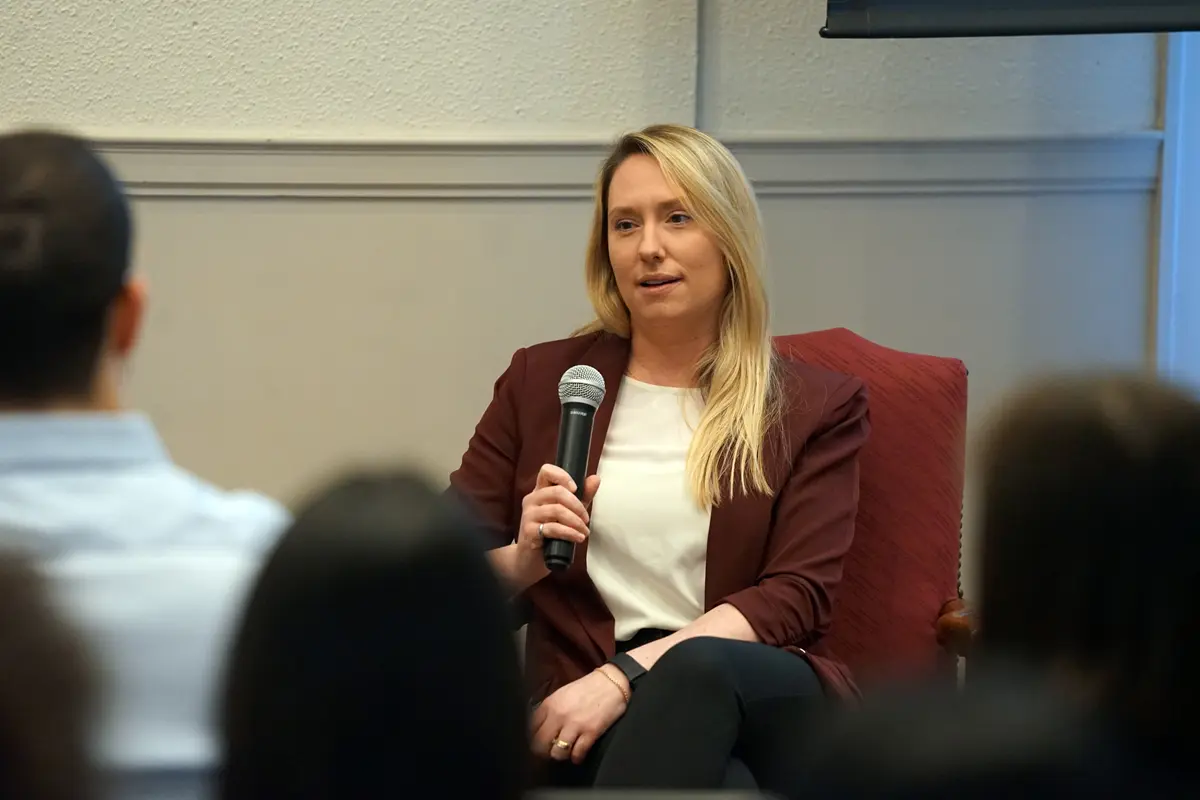 A woman sits in a chair and holds a microphone while speaking to people in a room.