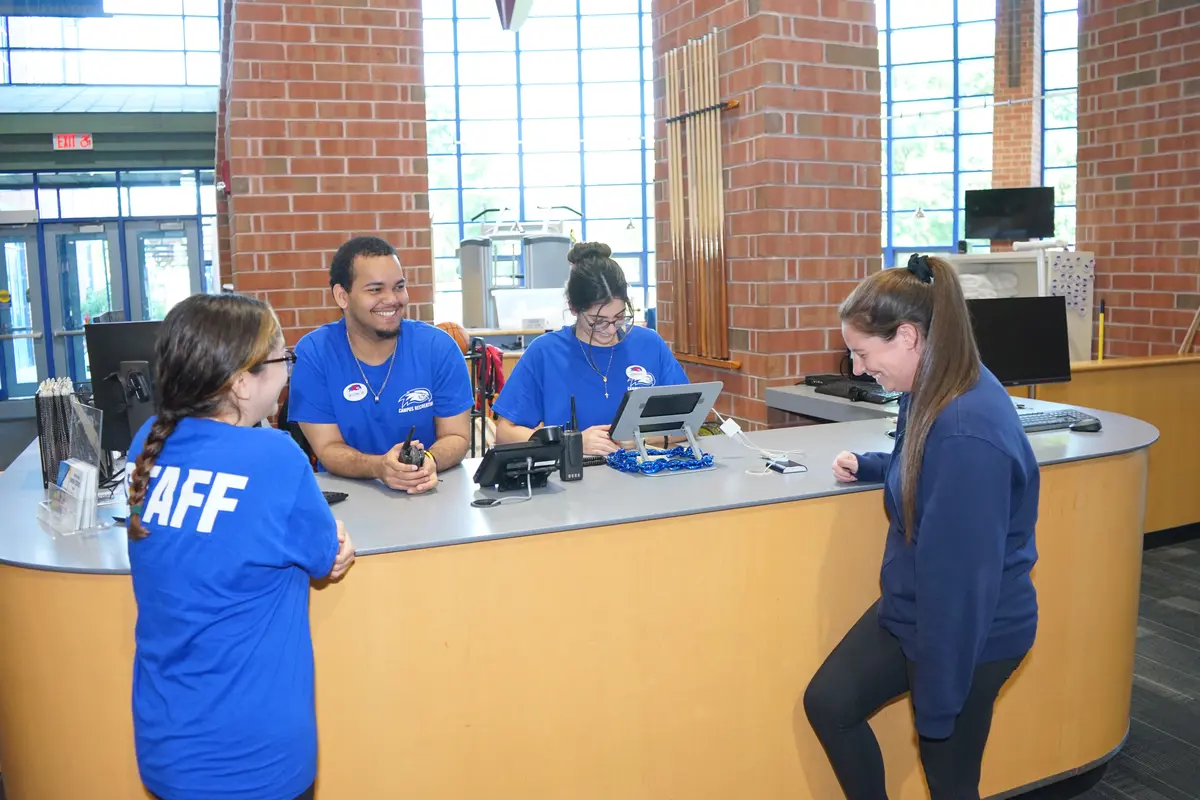 A woman laughs standing at the front desk of a campus rec center and talking to three student employees in blue T-shirts.