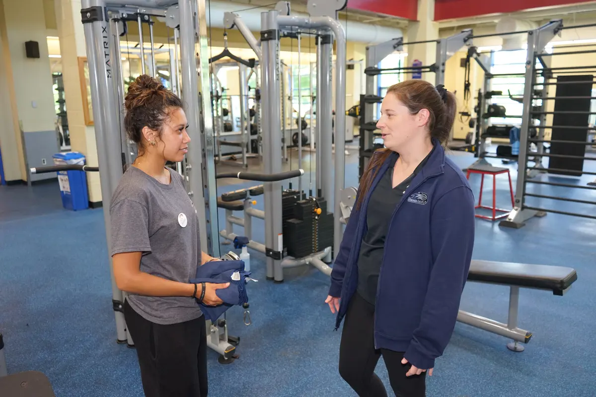 Two young women face each other and chat in a college campus recreation weight room.