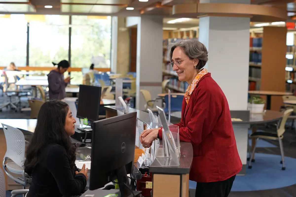 A woman in a red top stands at a reception desk and talks to a woman seated at a computer.