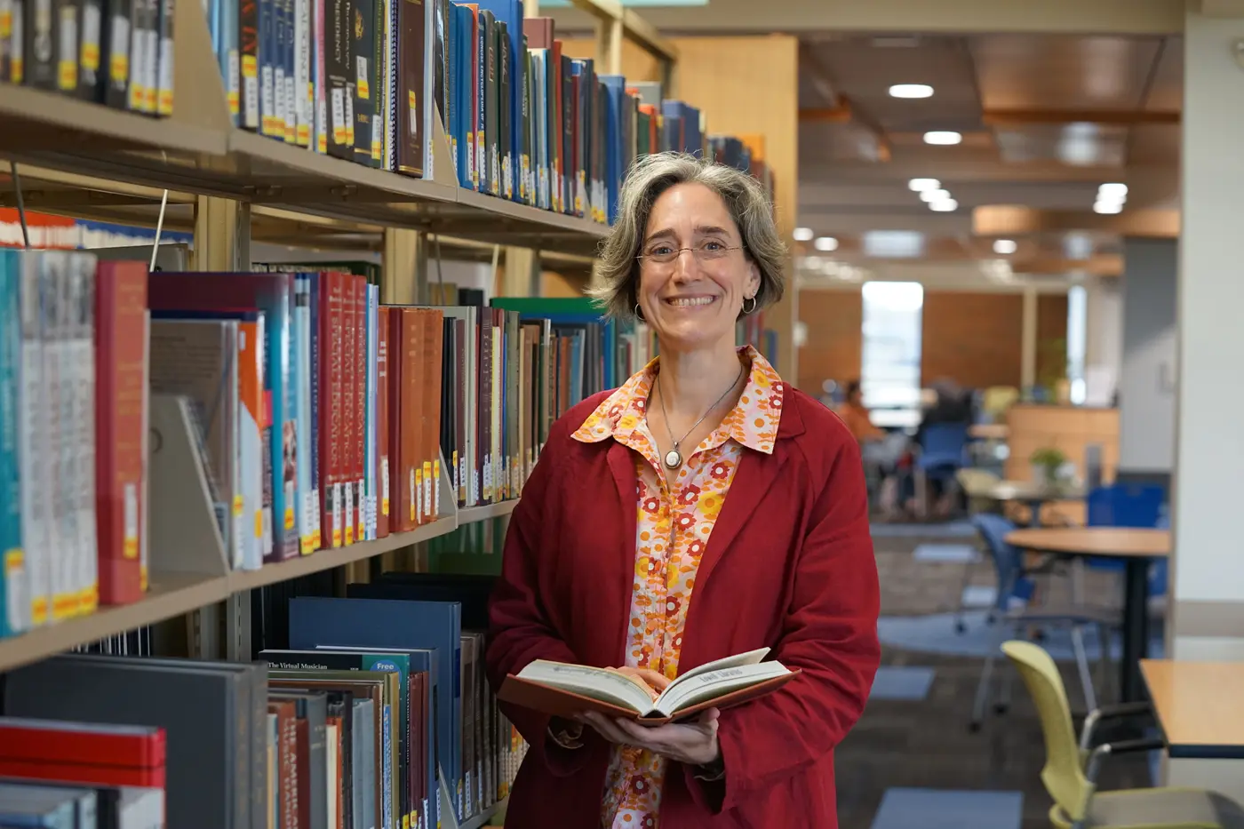 A woman smiles for a photo while holding a book near a shelf in a library.