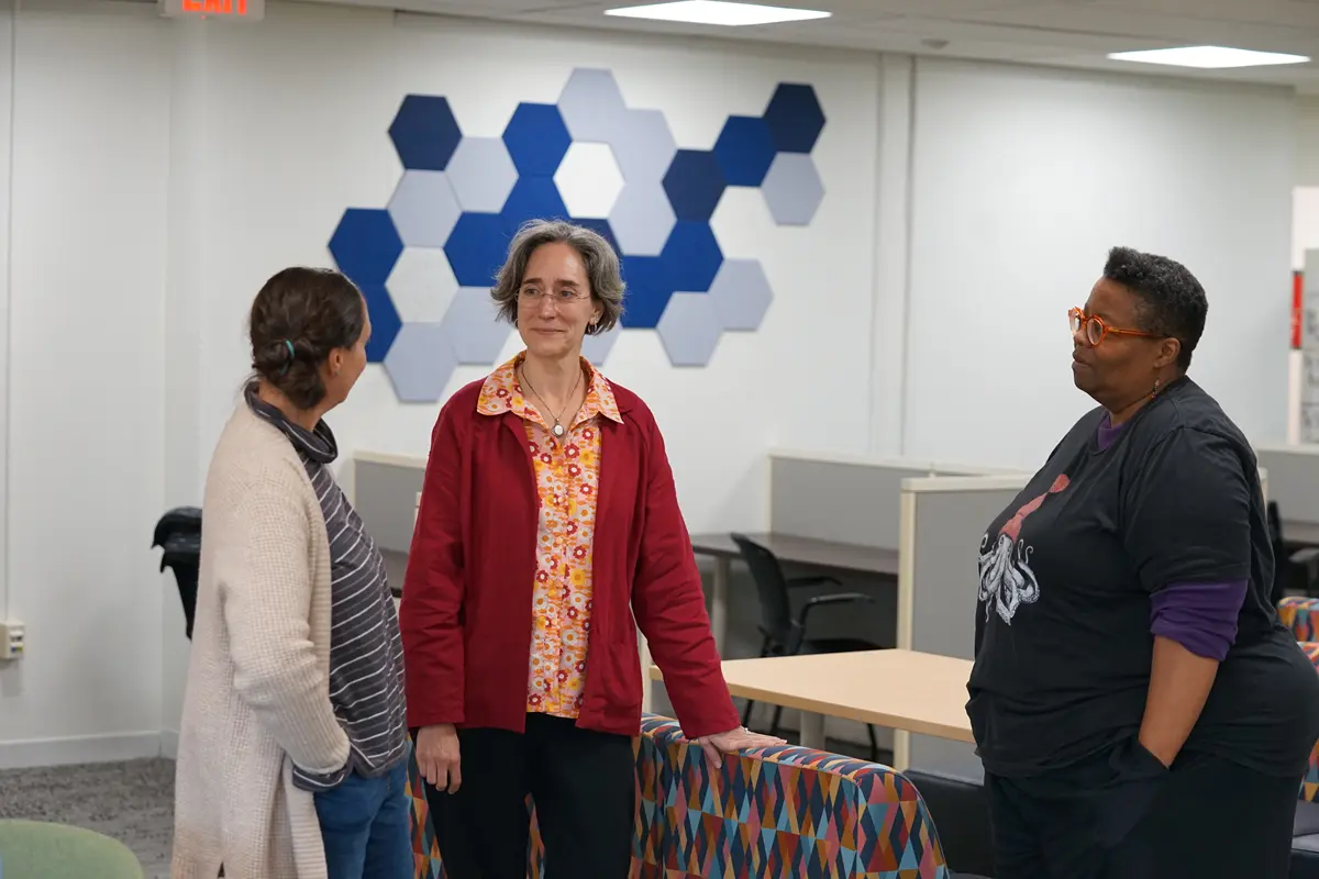 Three women stand in a study area and talk to each other.