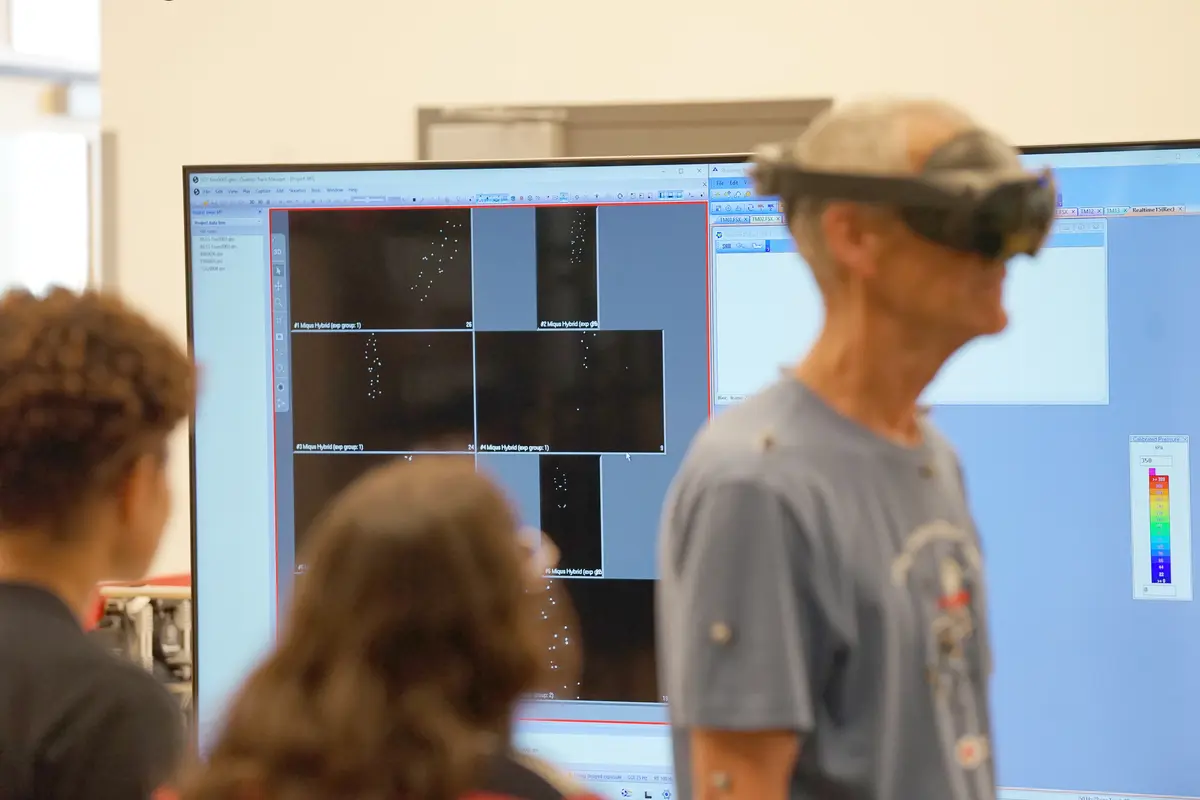 A man wears VR goggles in the foreground while a monitor in the background shows motion capture points.
