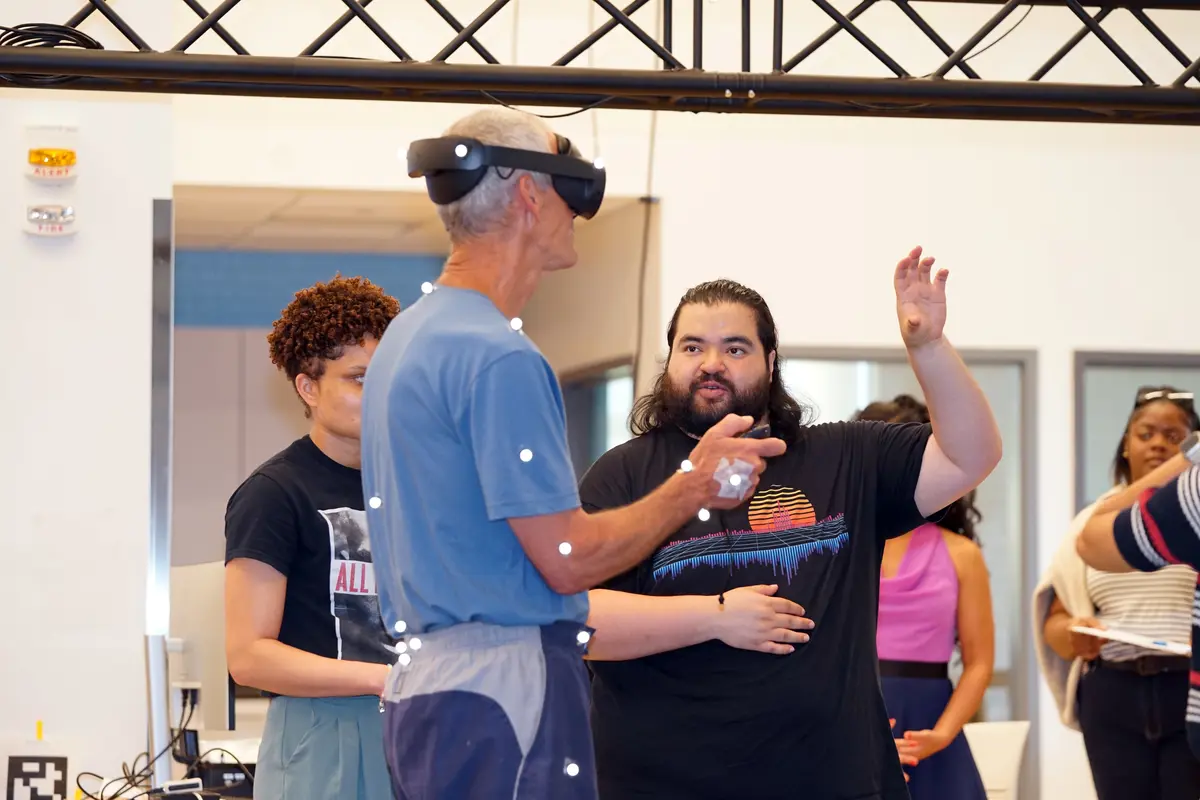 A young man with a beard gestures with his hand while talking to an older man who is wearing VR goggles and has sensors attached to his clothes in a lab.
