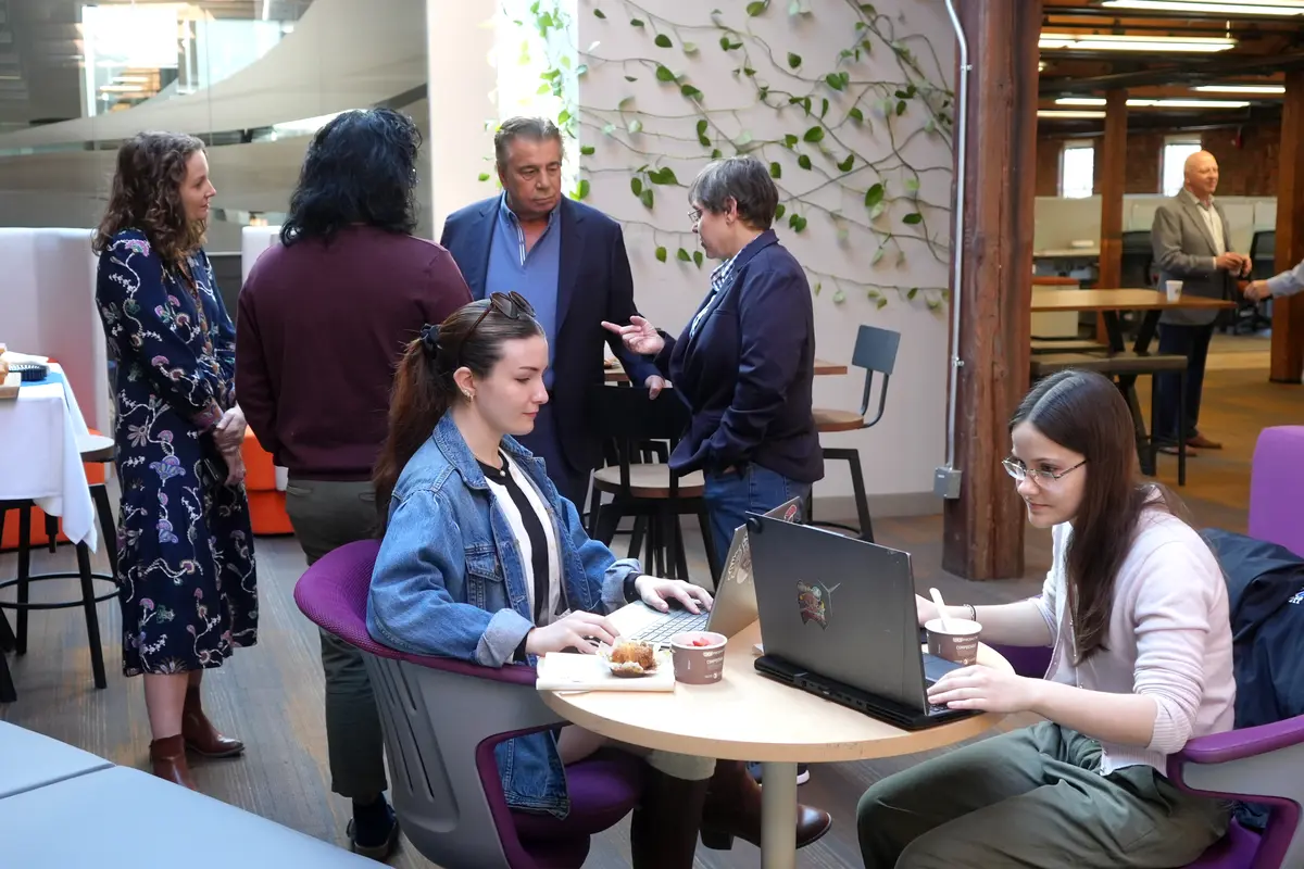 Two people work in foreground at table on laptop while four others talk standing in background
