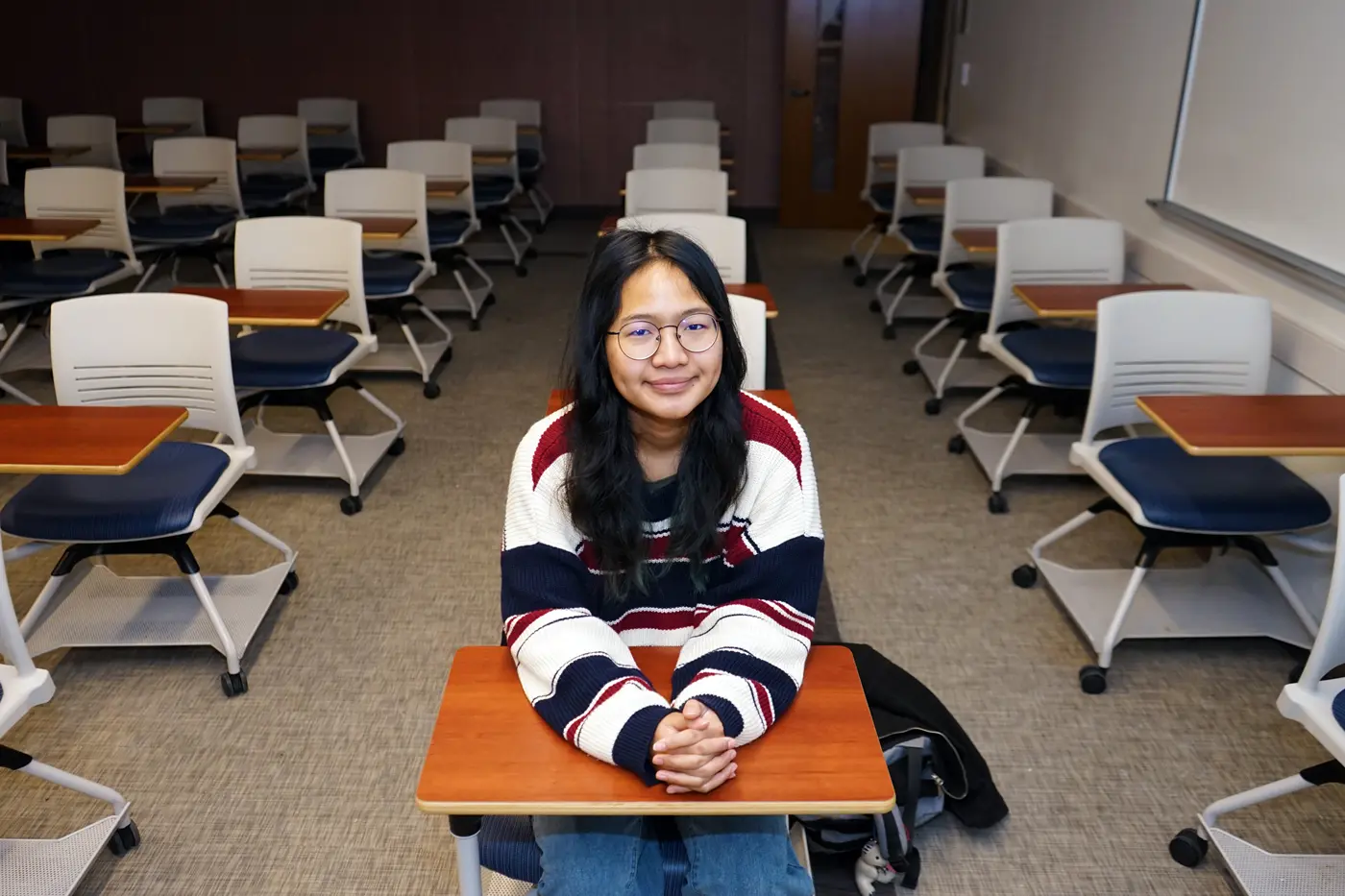 A student with glasses and dark hair poses for a photo while sitting at a desk in an empty classroom.