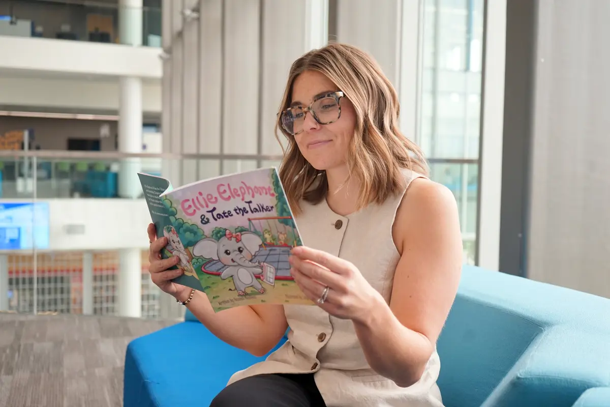 A young woman in glasses reads a childrens book while sitting on a blue couch by a window in a building.