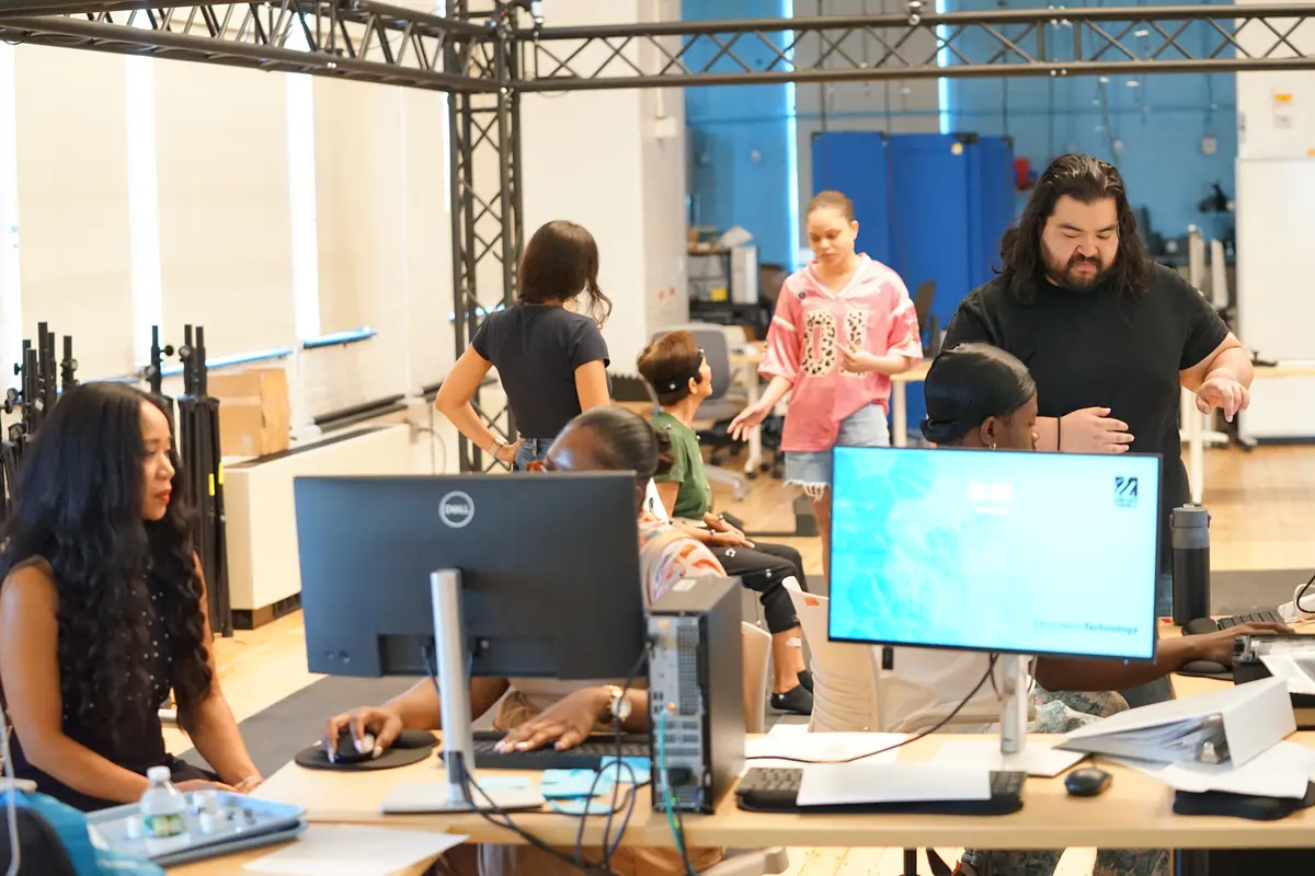 Four people work at computers at desks while two women talk to a woman who is seated in the background in a lab.