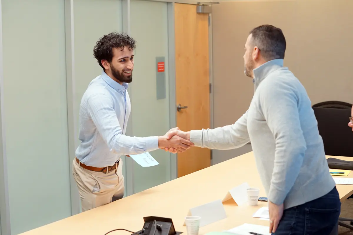A young man with a beard shakes hands with another man in a conference room.