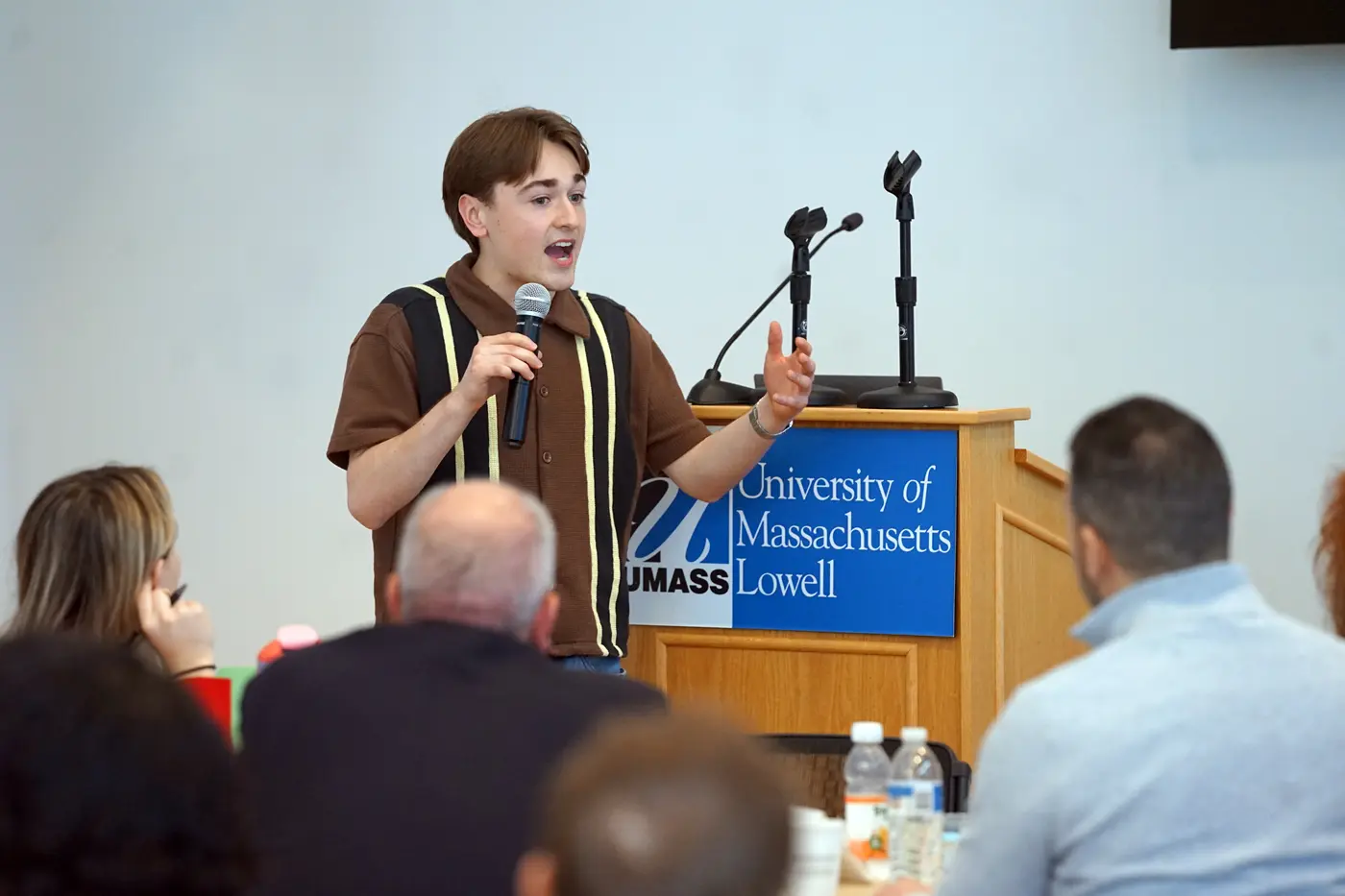 A young man in a striped shirt holds a microphone while taking to people seated in front of him.