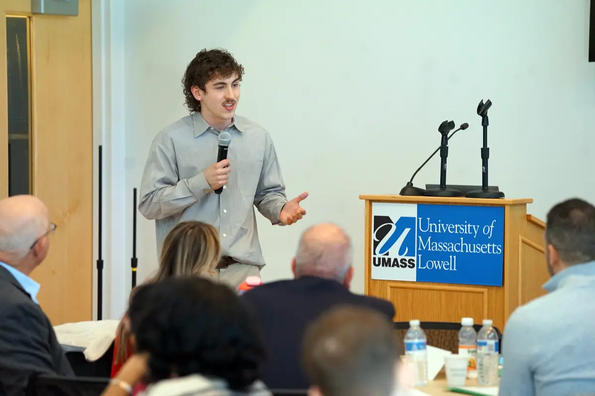 A young man holds a microphone and speaks to people sitting in a room.