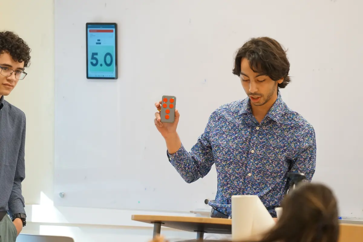 A student holds a clicker with red buttons in his left hand while sitting in a classroom.