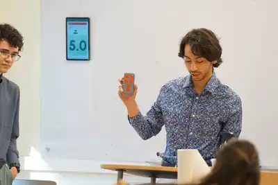 A student holds a clicker with red buttons in his left hand while sitting in a classroom.