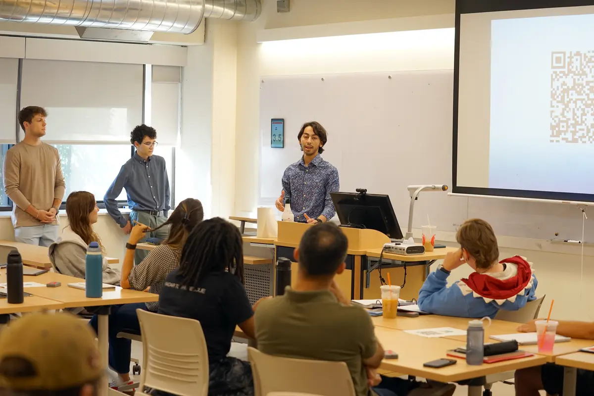 A young man in a blue shirt speaks to students in a class while two other young men look on from the side.
