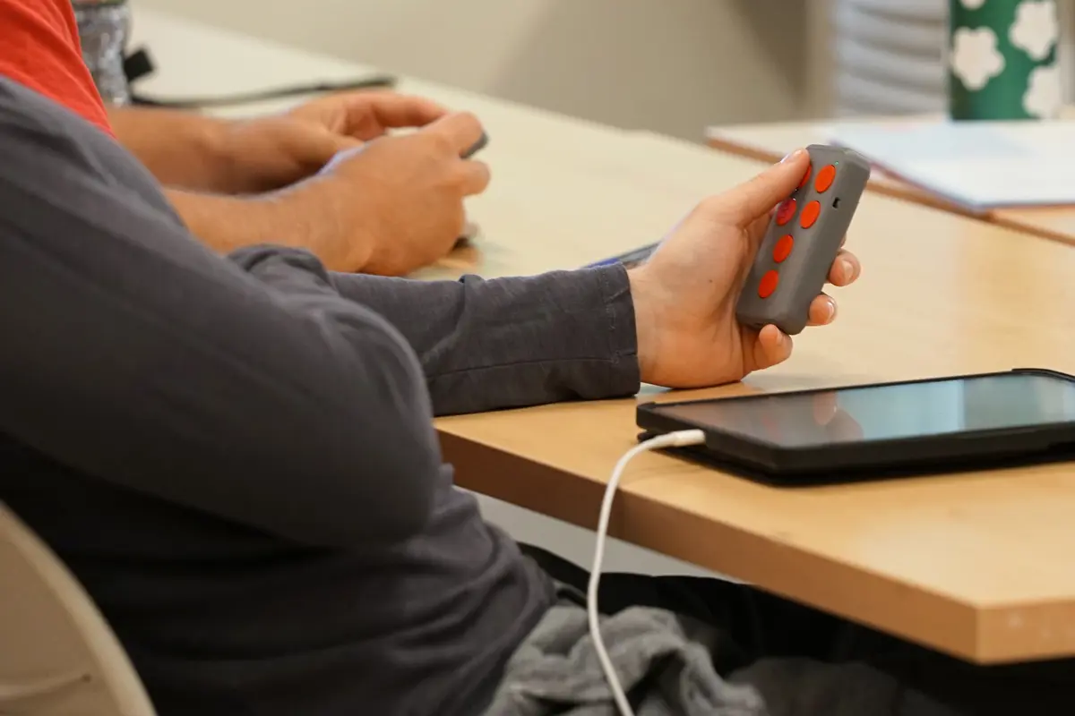 A student holds a clicker with red buttons in his left hand while sitting in a classroom.