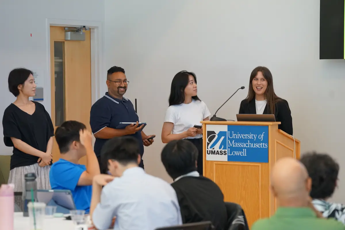 A young woman speaks at a podium while three people stand alongside her at the front of a room.
