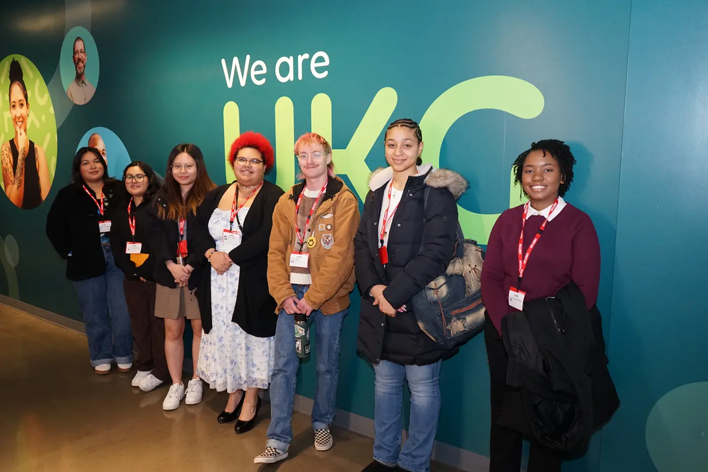 Seven college students pose for a photo while standing in front of a green wall.