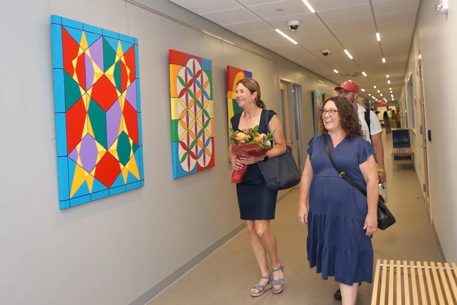 A woman holding a bouquet of flowers looks at artwork on a hallway wall while walking alongside two people.