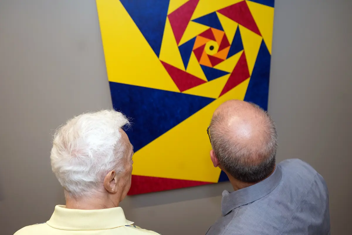 Two men look at a geometric painting hanging on a gray wall.