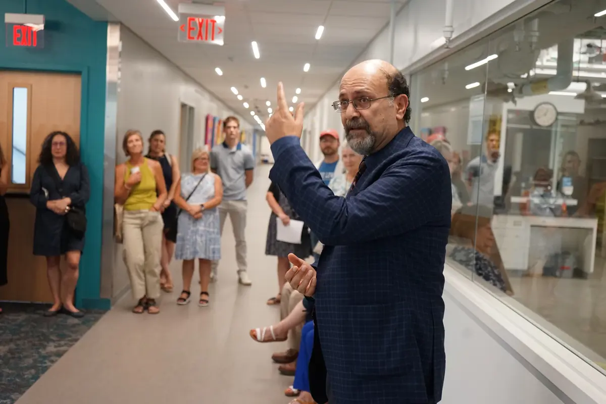A man in a blue blazer gestures with his left hand while speaking to people in a hallway.