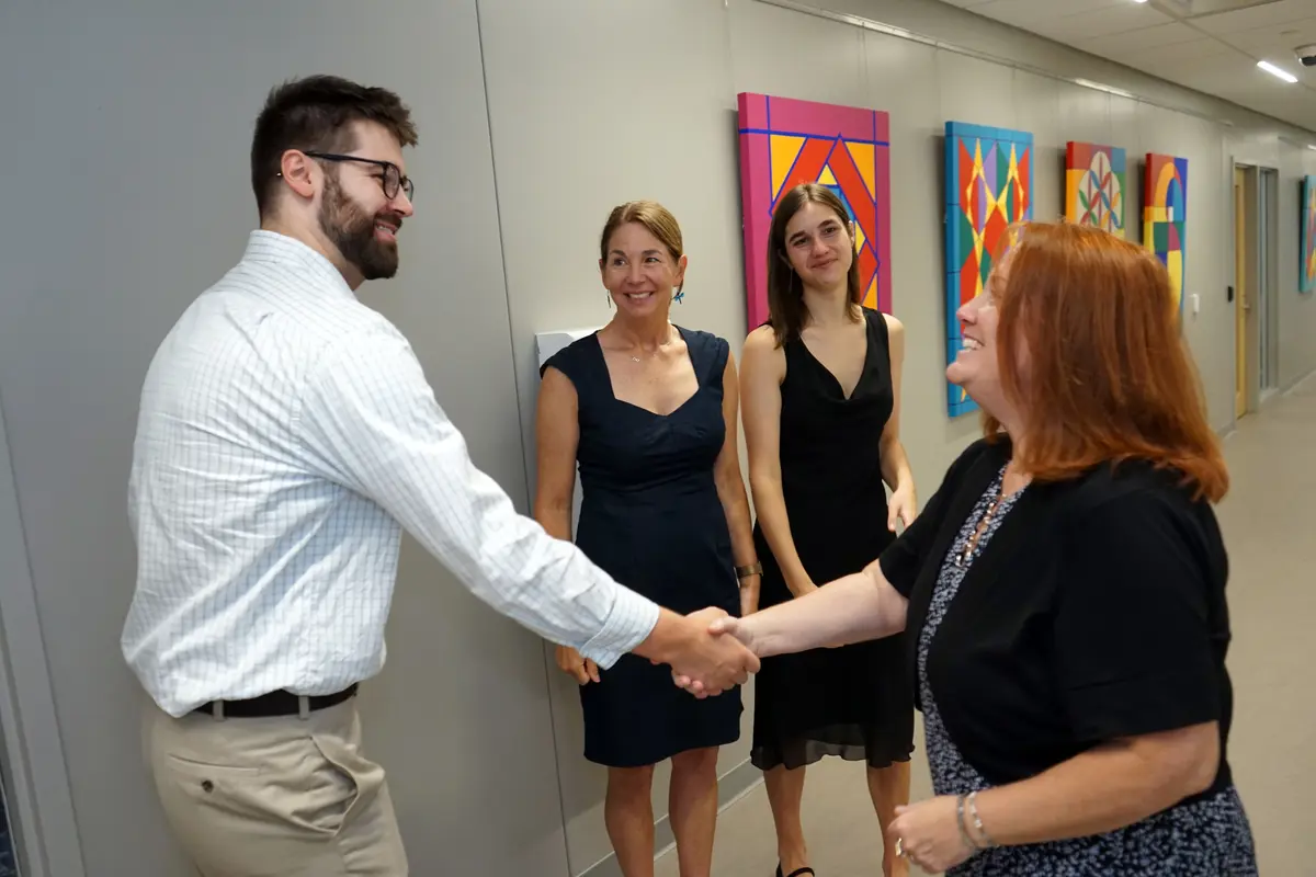 A man in a dress shirt shakes hands with a woman while two other women look on in a hallway.