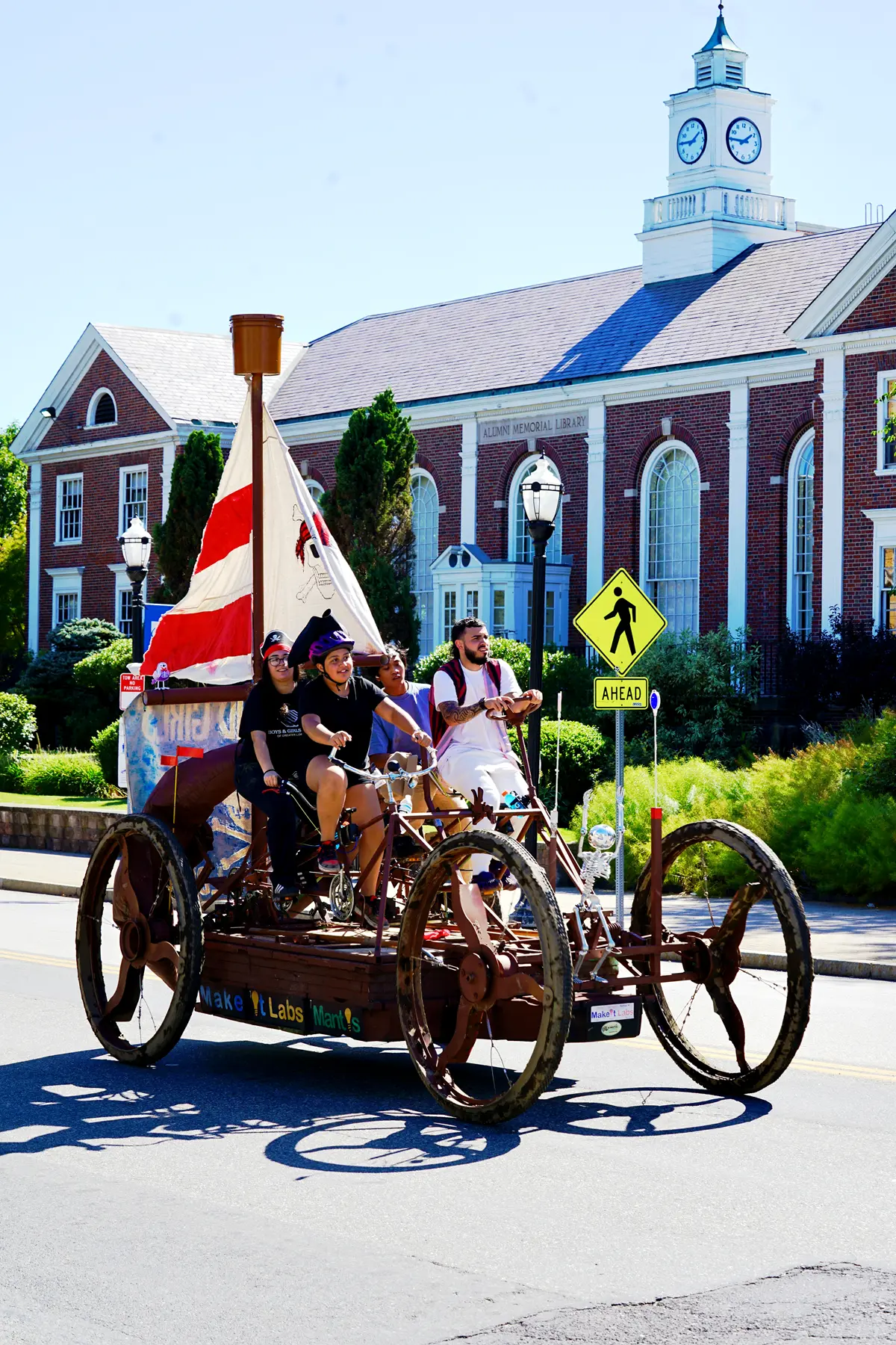 Four people on a bicycle powered pirate ship ride past a college building.