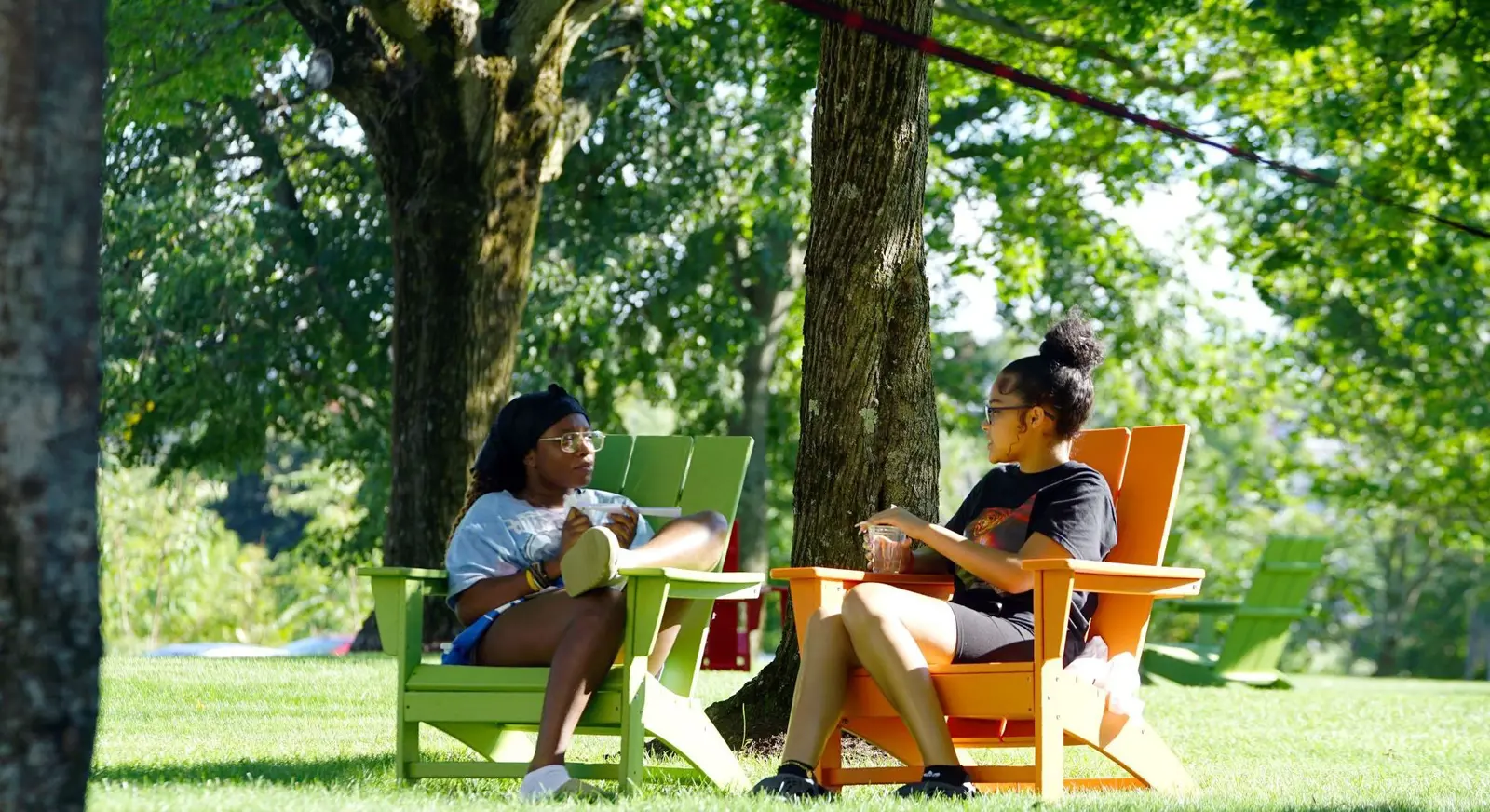 Two students sit in colorful Adirondack chairs under a tree