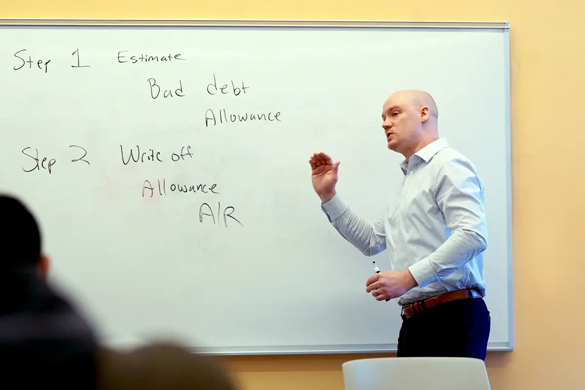 An accounting professor gesturs while standing in front of a dry erase board in a classroom.