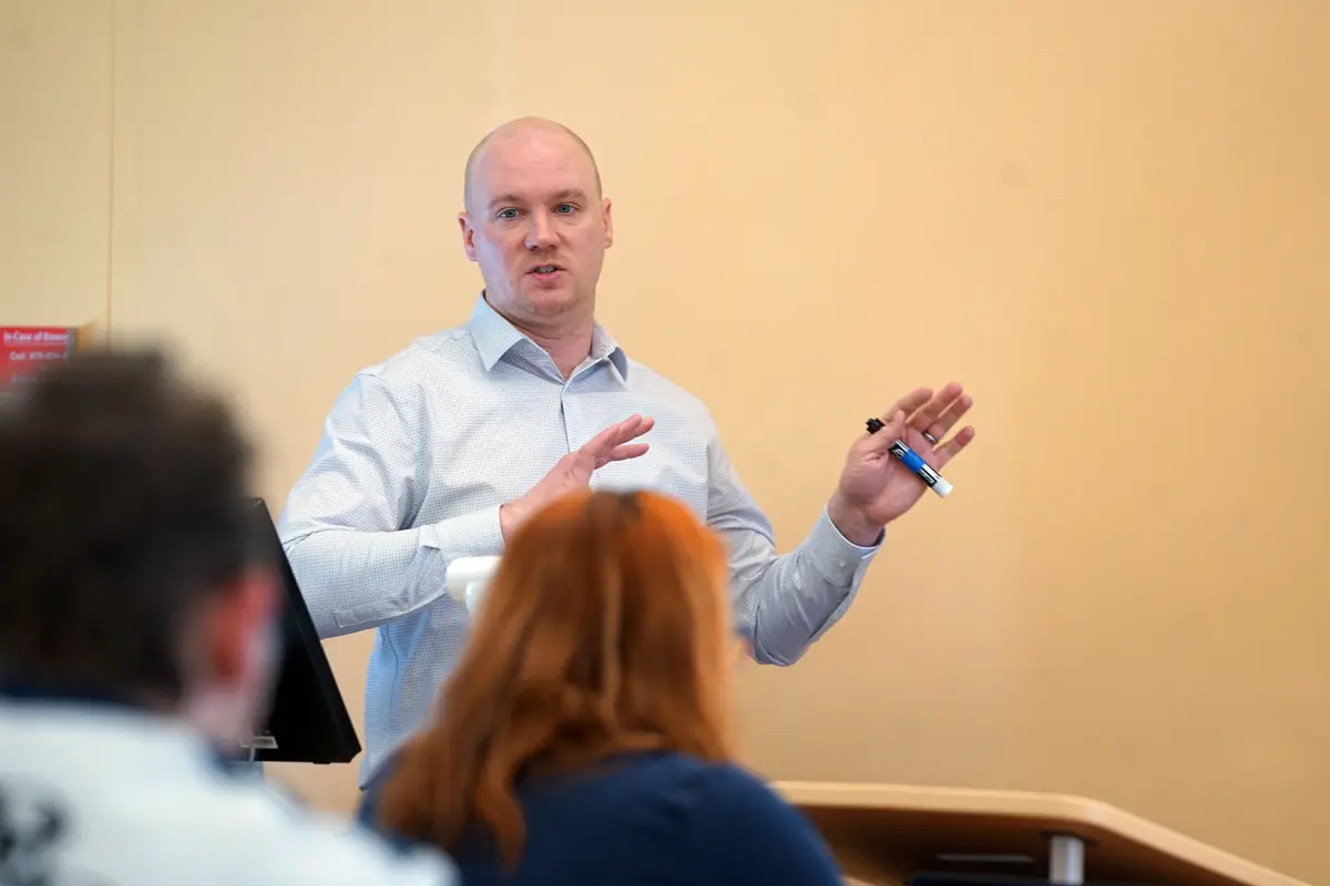 A man in a button-down shirt speaks in front of a class while holding a pen.