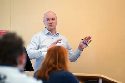 A man in a button-down shirt speaks in front of a class while holding a pen.