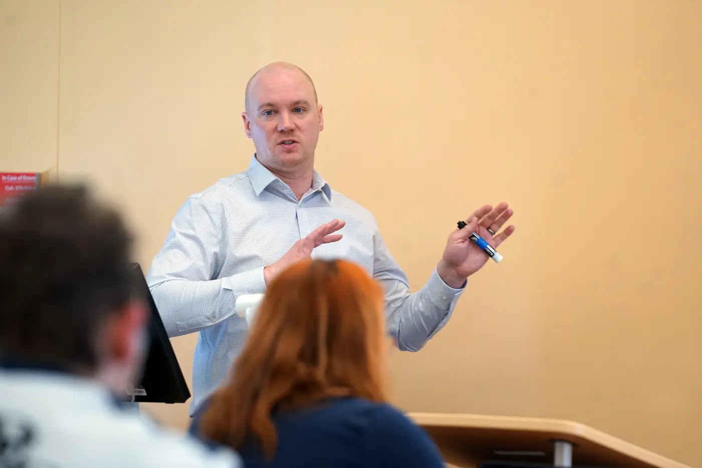 A man in a button-down shirt speaks in front of a class while holding a pen.