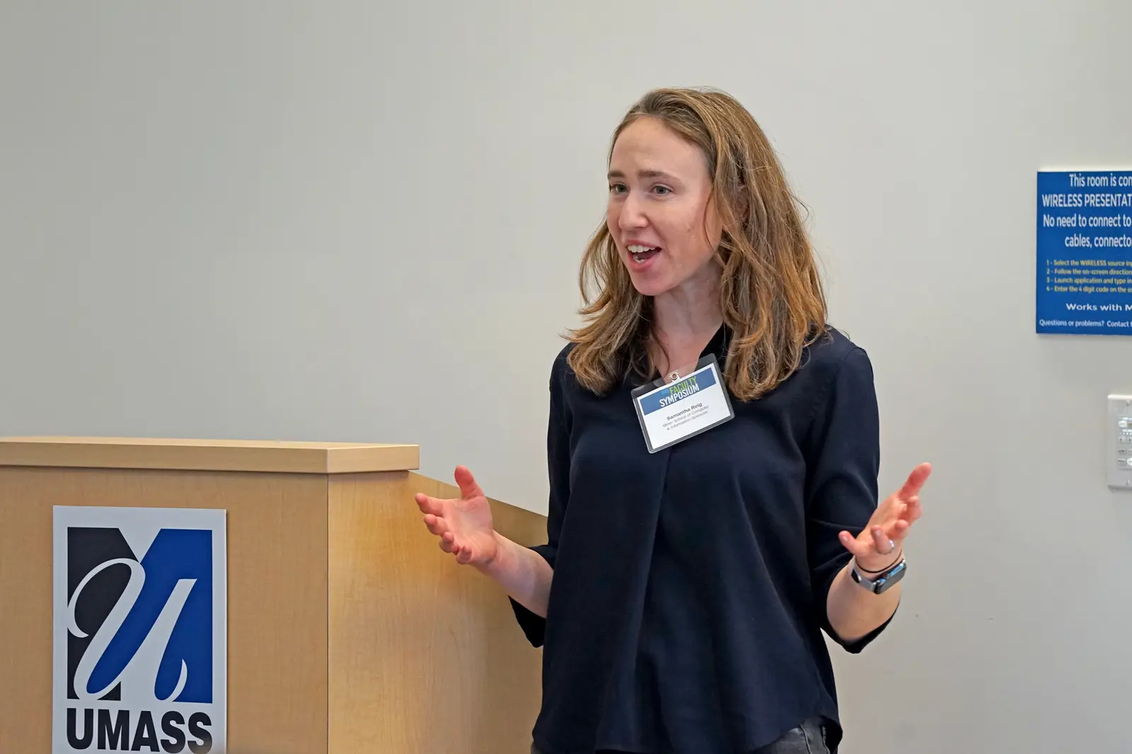 A woman gestures with her hands while giving a talk alongside a podium in a room.