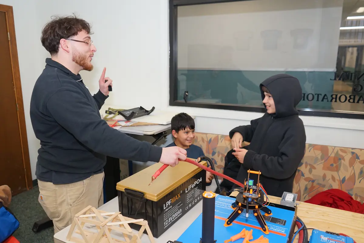 A young man in glasses waves his finger at two young boys in a college engineering lab.