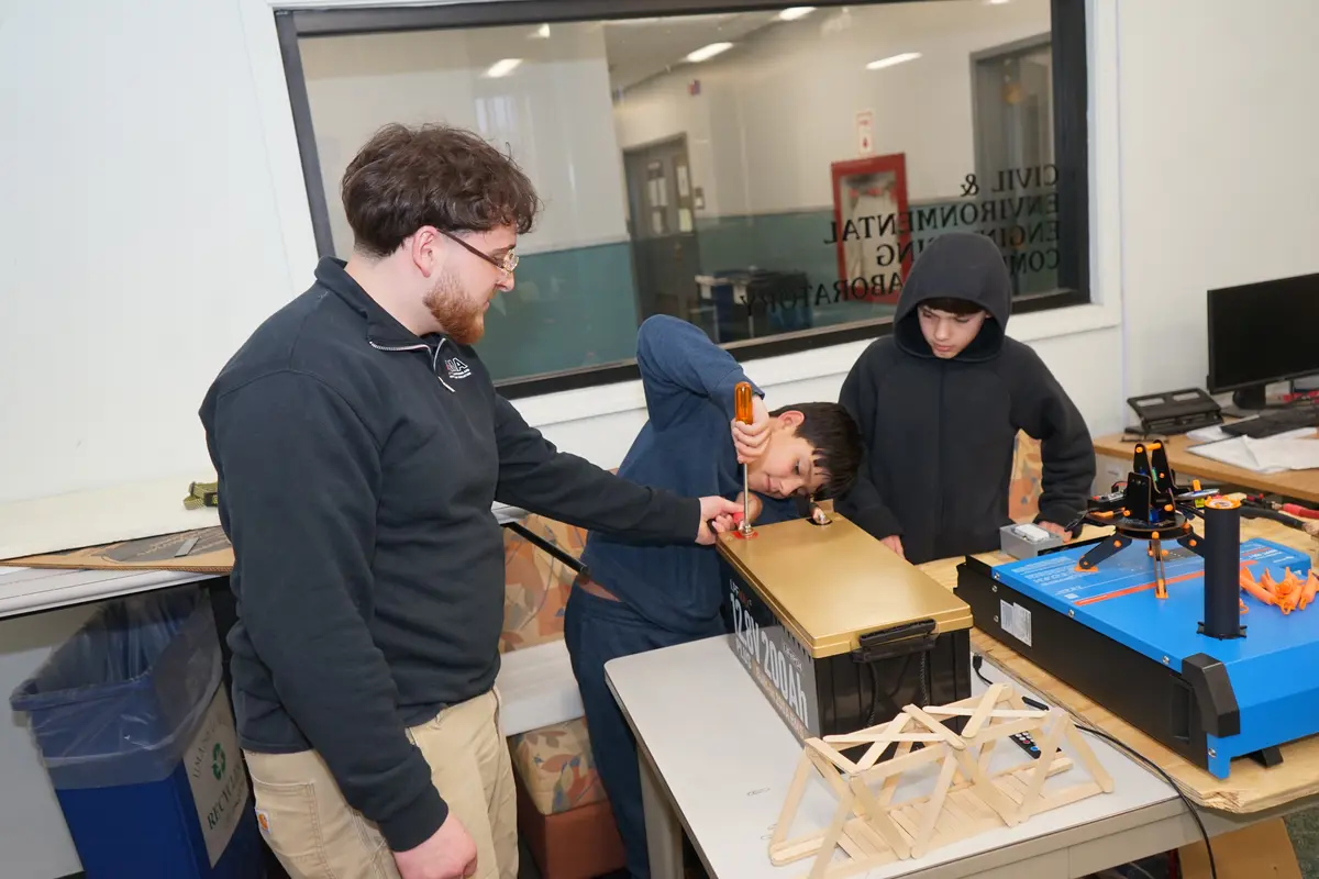 A boy uses a screwdriver on a battery while another boy in a hoodie and a college student look on in a room.