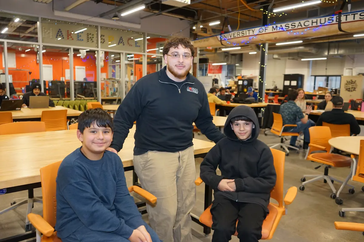 A young man in glasses poses for a photo with two young boys who are sitting in chairs in a college workshop.
