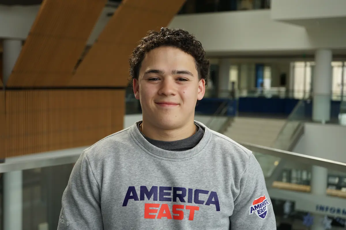 A young man with dark hair and weaering a gray sweatshirt poses for a photo in the lobby of a building.