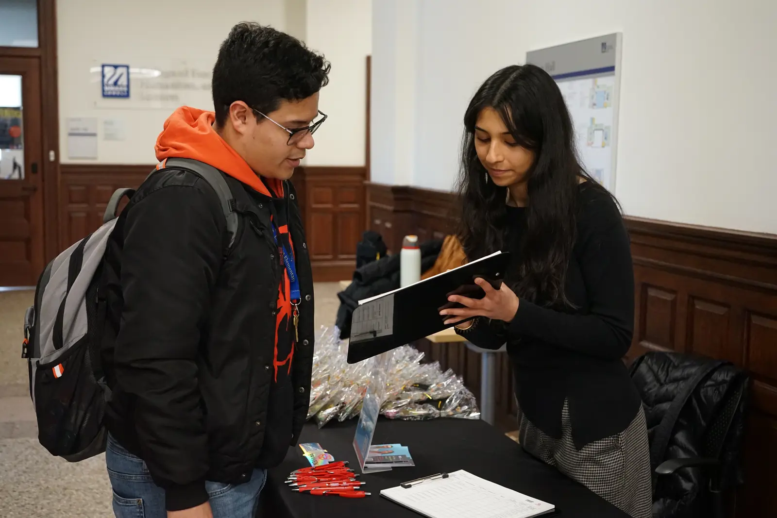 A young woman shows something on a clipboard to a young man wearing glasses and a backpack in a hallway.