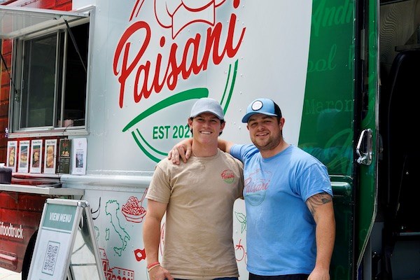 Two young men in T-shirts and caps pose for a photo in front of their food truck