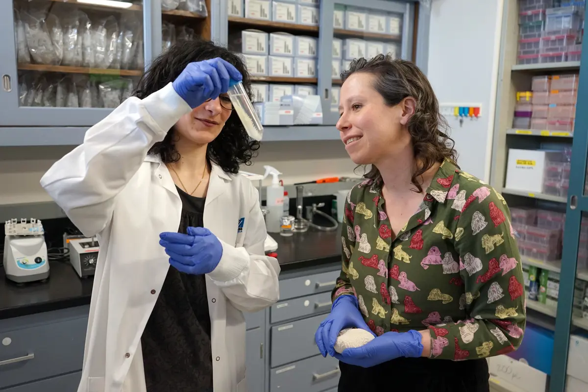 A young woman in a labcoat holds a glass beaker while a professor looks on.