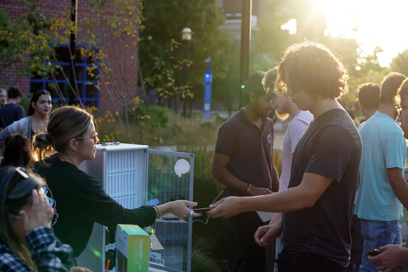 A young man hands his phone to a young woman at a table at an outdoor event at sunset.