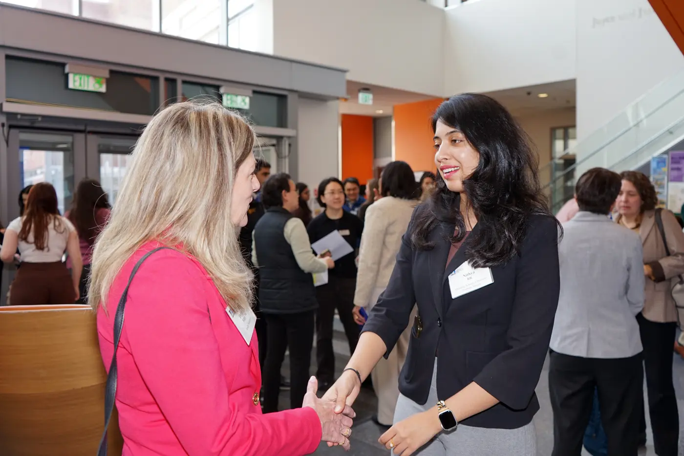 Two women in business attire shake hands in the crowded lobby of a building.