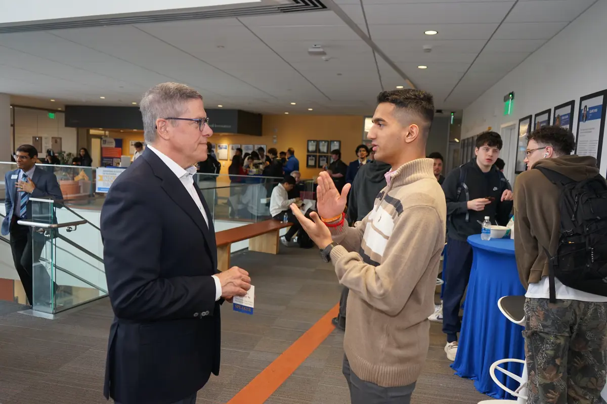 A young man gestures with his hands while speaking with a man wearing a suit in a crowded building foyer.