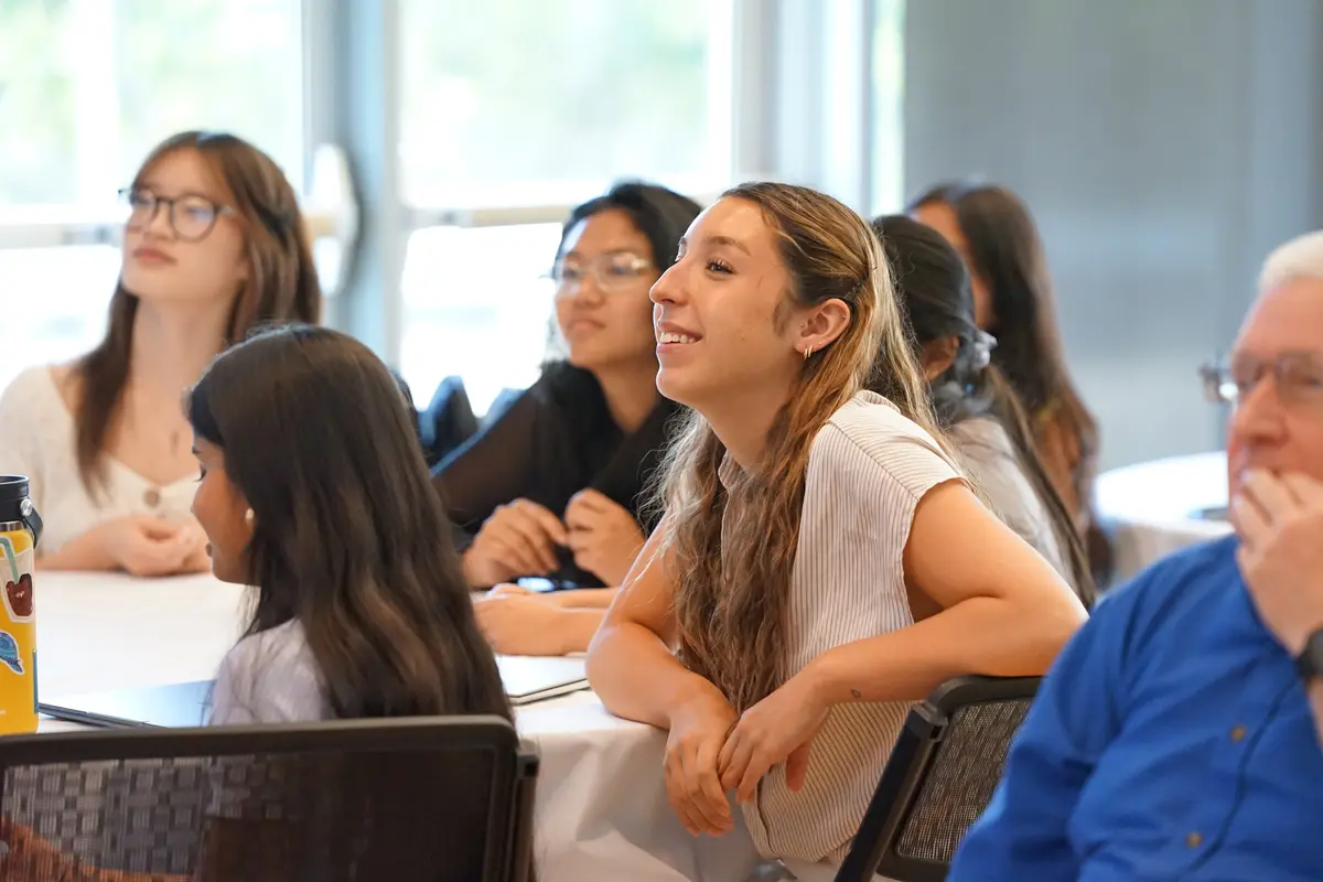 A young woman smiles while listening to a presentation while seated at a table with other people.