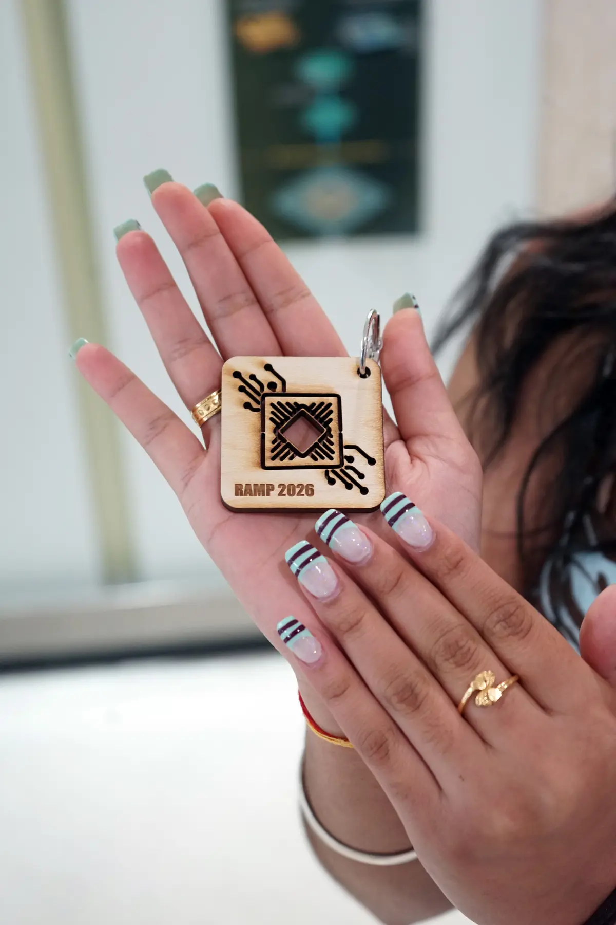 A young woman with painted nails holds a wooden keychain that has the RAMP 2026 logo on it.