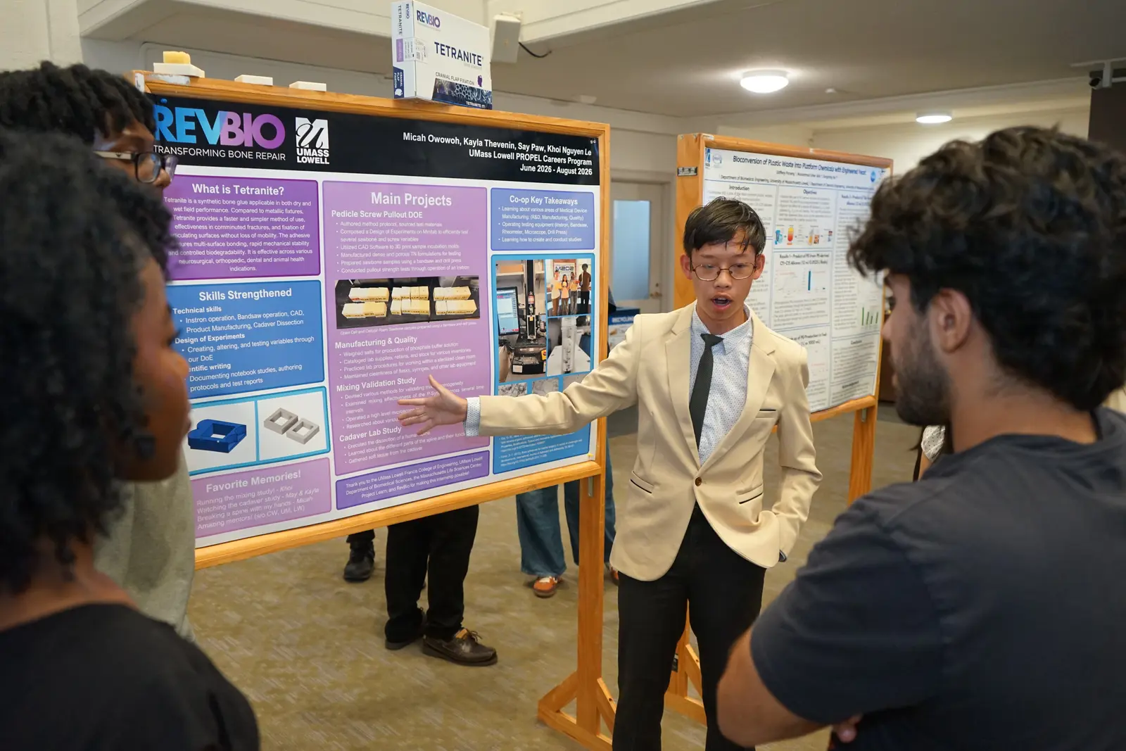 A young man in a tie and tan blazer gestures to a research poster while talking to people in a room.