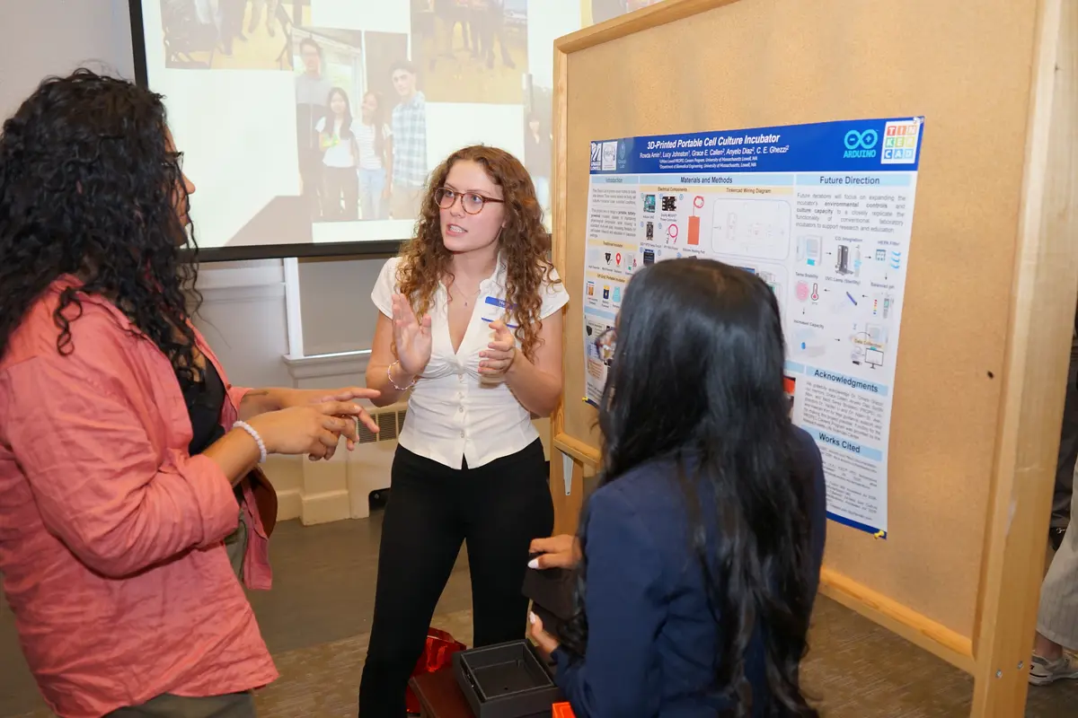 Three young women talk in front of a research poster and projection screen.