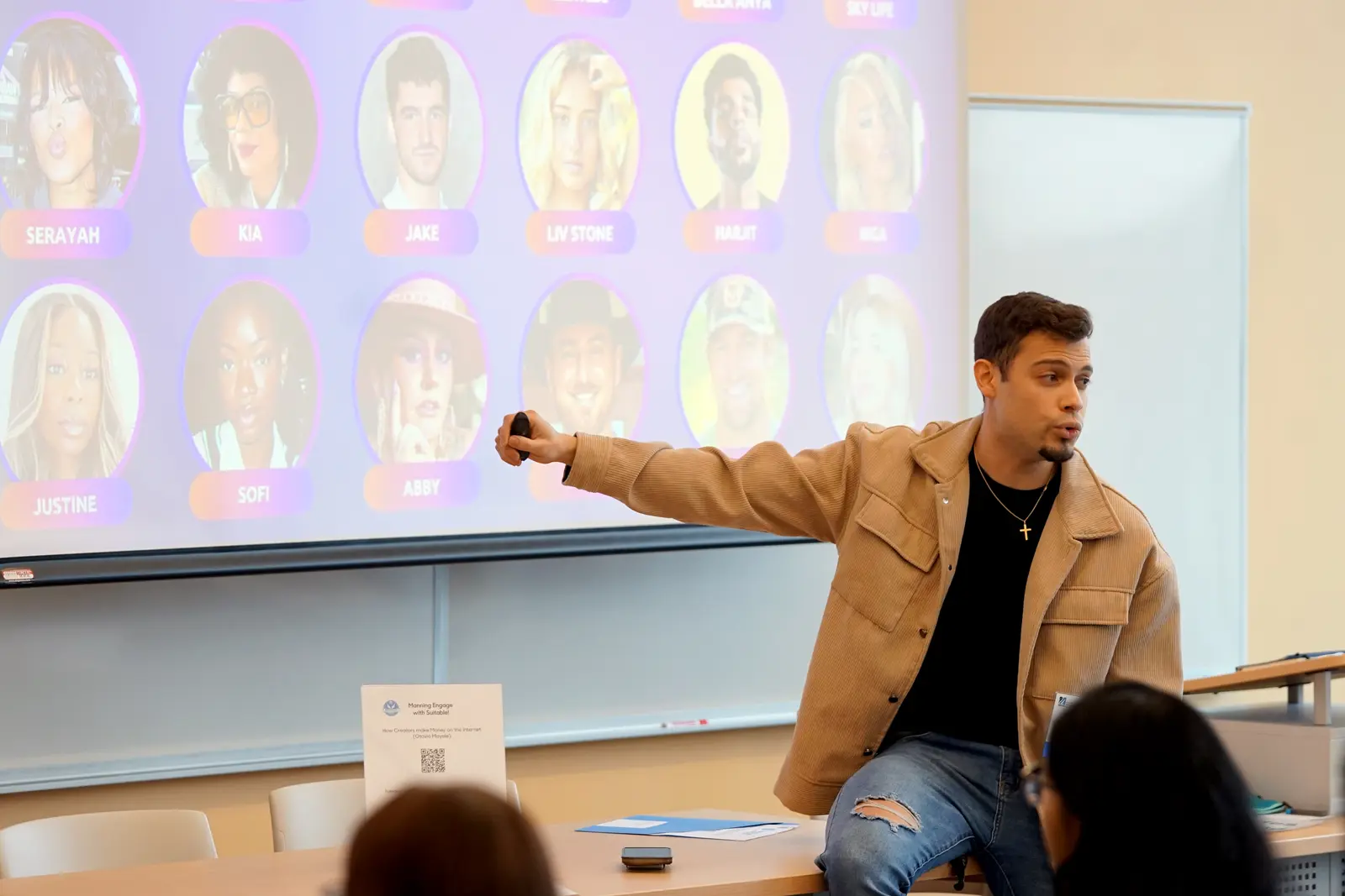 A young man in a tan jacket points to a projection screen that has images of people's faces while speaking to a classroom.